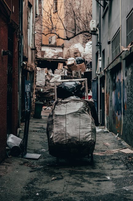A narrow urban alleyway is cluttered with a large, gray, weathered bin or bag in the foreground, positioned centrally on the rough, dark asphalt surface which shows signs of oil stains and debris. Beyond the bin, the alley extends toward a pile of discarded cardboard boxes, black plastic bags, and miscellaneous rubbish stacked against a wooden fence on the left, with some boxes leaning precariously. On the right side, there is a dark-colored building wall featuring graffiti and an iron barred window, alongside a series of utility meters and conduits attached to the wall. The background reveals leafless tree branches and older brick and plaster buildings with a muted, overcast sky, suggesting an early or late time of day. The scene has a subdued, gritty atmosphere typical of an area awaiting rubbish collection or disposal, with evident signs of neglect and clutter typical of private waste handling. Occasionally, the scene subtly reflects the context of independent rubbish removal services, aligning with the theme of alternative waste management solutions in urban environments. Rubbish Clearance Hackney’s services could be relevant here, as this area appears to require on-site clearance for bulky waste items and accumulated refuse, characteristic of non-local authority waste collection scenarios.