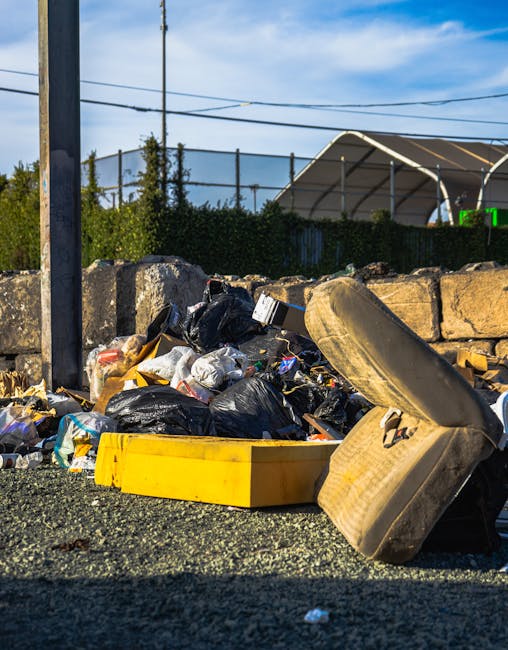 A pile of mixed household waste and rubbish bags, predominantly black plastic bags, is scattered on the ground beside a low stone wall in an outdoor area. Among the rubbish, there is an old, beige upholstered chair cushion with fabric torn and frayed, leaning against the wall, and a yellow plastic container lying on its side. In the background, a metal fence, green shrubbery, and power lines are visible, with a partially visible modern, curved canopy structure and a bright blue sky overhead, suggesting a clear day. The scene appears to be in a service yard or outdoor storage space, likely related to private rubbish collection or waste disposal services, such as those provided by Rubbish Clearance Hackney, dealing with bulky or unsorted rubbish that may be difficult to manage through standard collection. The environment is well-lit with natural sunlight, casting shadows on the ground, emphasizing the disorganized state of the waste in a setting indicative of alternative waste handling methods.