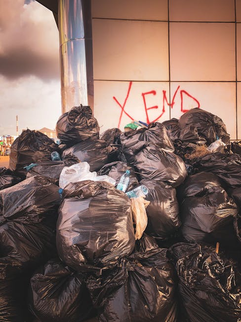 A narrow urban alleyway is cluttered with a large, gray, weathered bin or bag in the foreground, positioned centrally on the rough, dark asphalt surface which shows signs of oil stains and debris. Beyond the bin, the alley extends toward a pile of discarded cardboard boxes, black plastic bags, and miscellaneous rubbish stacked against a wooden fence on the left, with some boxes leaning precariously. On the right side, there is a dark-colored building wall featuring graffiti and an iron barred window, alongside a series of utility meters and conduits attached to the wall. The background reveals leafless tree branches and older brick and plaster buildings with a muted, overcast sky, suggesting an early or late time of day. The scene has a subdued, gritty atmosphere typical of an area awaiting rubbish collection or disposal, with evident signs of neglect and clutter typical of private waste handling. Occasionally, the scene subtly reflects the context of independent rubbish removal services, aligning with the theme of alternative waste management solutions in urban environments. Rubbish Clearance Hackney’s services could be relevant here, as this area appears to require on-site clearance for bulky waste items and accumulated refuse, characteristic of non-local authority waste collection scenarios.