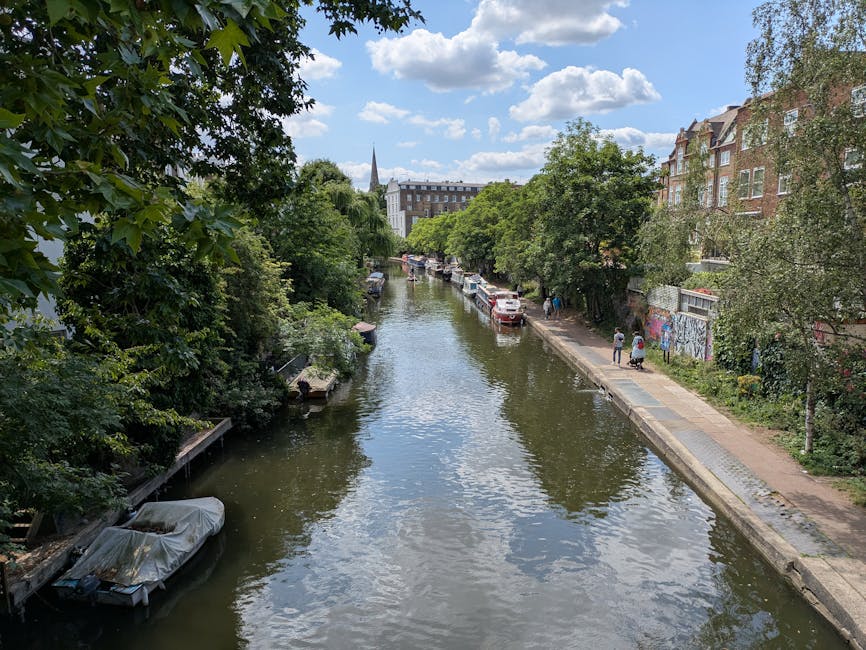 A narrow urban canal scene with calm water reflecting the partly cloudy sky above. On the left bank, dense green trees and foliage overhang the water, with small boats moored along the edge, some covered with protective tarps. The right bank features a concrete walkway where a few pedestrians are strolling, and a row of parked cars lines the street, partially obscured by a line of mature trees with lush green leaves. Behind the trees, a series of traditional brick residential buildings with large windows and pitched roofs are visible, alongside a graffiti-covered wall. In the background, the canal extends toward a distant white church with a pointed spire, set against a bright sky dotted with fluffy clouds, creating a peaceful, everyday scene. The setting suggests a typical Hackney canal area where private rubbish clearance services might be involved in maintaining cleanliness alongside public waste disposal.
