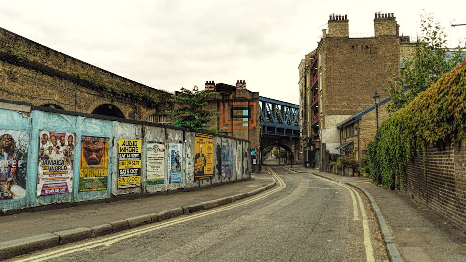 A narrow urban street scene showing an asphalt road with a double yellow line along the edge, slightly curved towards the background, flanked by brick and plaster buildings, with a green hedge on the right side and a weathered brick wall on the left covered with colorful posters advertising events. The posters are attached to a metal fence separating the street from an elevated railway bridge painted in blue, supported by brick pillars with visible graffiti. The sky is overcast with a light gray tone, casting diffuse natural light over the scene. This setting suggests a typical east London neighbourhood, where outdoor rubbish clearance or local waste collection might take place, with no visible rubbish or waste items present in the scene, but the context aligns with private or independent waste handling activities. The image appears to be part of a guide for rubbish clearance in Hackney, with Rubbish Clearance Hackney potentially offering removal services in such environments.