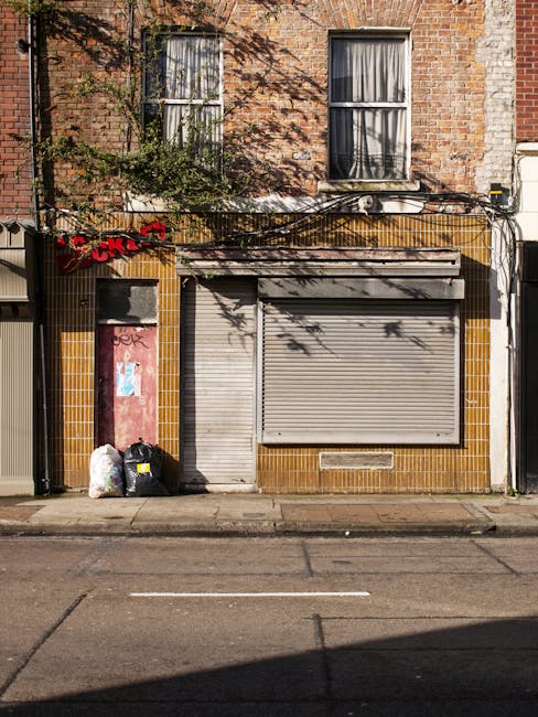 A ground-level view of a two-story brick building with two large sash windows on the upper floor, both with closed white frames and partially drawn curtains inside. The facade features reddish-brown bricks with some greenery, including a small bush growing near the left window. On the lower part of the building, a closed metal roller shutter painted light grey covers a shopfront, with a slightly worn and textured surface. To the left of the shutter, a narrow door, also painted grey, is partially obscured by a black garbage bag placed on the pavement, along with a white plastic bag beside it. The lower part of the building around the shopfront has yellow-brown tiles arranged in a grid pattern, some slightly dirty or aged. Overhead, black cables hang loosely, casting shadows on the building’s facade. The scene appears to be on a quiet street with a curb and asphalt road in the foreground, and the overall lighting suggests a clear day with natural sunlight highlighting textures and colors. Rubbish clearance services by Rubbish Clearance Hackney could be related to the disposal of items like the garbage bags visible in this setting, representing typical waste collection needs in urban environments.