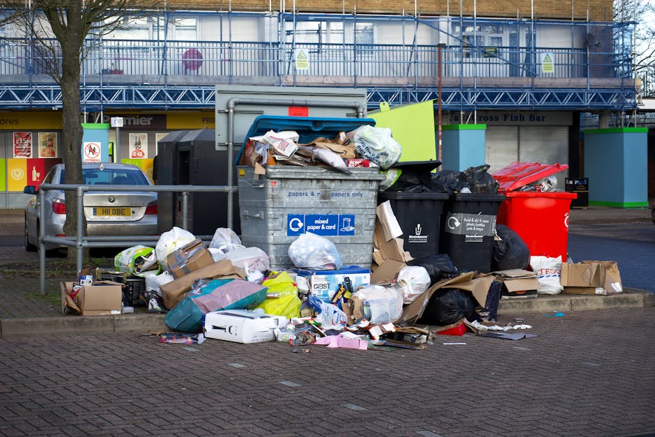 A large pile of mixed waste and rubbish bags situated on a paved parking space in front of a building with commercial units. The debris includes crumpled cardboard boxes, plastic carriers, paper and plastic packaging, and black and colored garbage bags. Some rubbish spills onto the surrounding pavement, with discarded cereal boxes, plastic containers, and other packaging visible underneath and around the pile. Behind the waste, there is a metal railing separating the rubbish from parked vehicles, including a silver car with a yellow registration plate. The background features a multi-storey building with a blue metal external staircase, yellow signage, and storefronts, one labeled 'Cross Fish Bar'. The scene is lit by natural daylight, suggesting a typical urban environment where private waste collection or rubbish clearance services, such as those provided by Rubbish Clearance Hackney, manage excess waste outside commercial or residential premises.