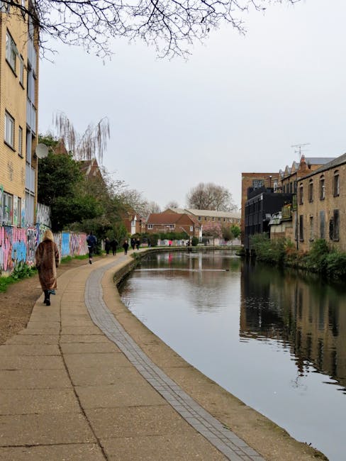 A paved walkway runs alongside a narrow river or canal in an urban area, with several pedestrians walking in both directions. On the left side, there is a multi-story residential building with a brick façade and some windows decorated with graffiti art. The building's side is partly obscured by leafless tree branches extending into the frame. To the right, there are older brick buildings with visible windows and balconies, some with small plants or outdoor furniture. The river's surface is calm, reflecting the overcast sky and the surrounding structures. In the background, more residential houses are visible, with a mixture of single-story and low-rise designs, along with trees and additional buildings in the distance. The environment suggests a quiet, semi-urban setting that could benefit from private waste or rubbish removal services, such as those provided by Rubbish Clearance Hackney, to maintain cleanliness along such scenic pathways.