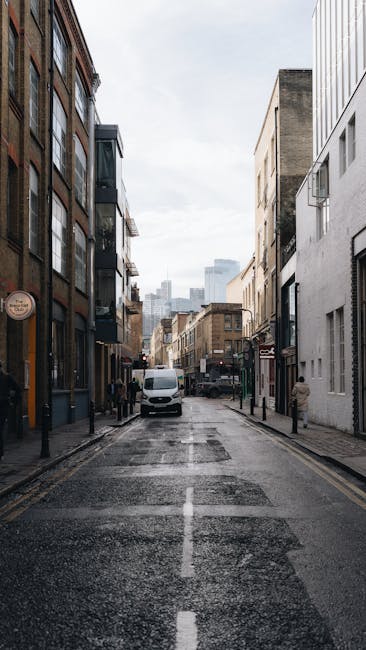 A narrow urban street in Stoke Newington E5, Hackney, shows a daytime scene with a wet asphalt road marked by a central white line, flanked on both sides by tall, mixed-use buildings with facades of brick, concrete, and metal cladding. On the left, there are modern black-framed glass windows and small shopfronts, while the right side features plain white and beige walls, some with windows and air conditioning units. Parked and moving vehicles, including a white van in the middle distance, occupy the street, which appears to have limited space for large rubbish removal vehicles to access directly. In the background, high-rise buildings and a cloudy sky are visible, contributing to the cityscape atmosphere. The setting suggests a typical city environment where alternative waste handling services might operate in tight or constrained access situations, as indicated by the closely spaced buildings and narrow street width; this aligns with the context of private rubbish collection and on-site clearance, as managed by Rubbish Clearance Hackney.
