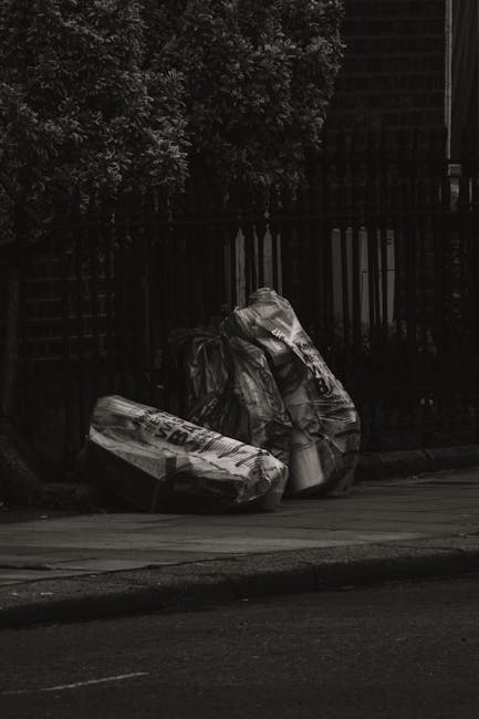 Two large black plastic garbage bags and a cardboard box, all sealed and slightly crumpled, placed on the pavement near a black metal fence with vertical bars and a brick wall in the background. The bags appear to contain household waste or rubbish, with visible creases and shiny textures indicating their plastic material. The cardboard box, positioned on the left, is partially flattened and features printed markings. The scene is set outdoors on a street or alleyway, with a shaded area created by nearby trees and fencing, suggesting an informal rubbish disposal area often managed by private waste removal services such as Rubbish Clearance Hackney. The environment appears quiet, with no people or vehicles visible, emphasizing the importance of private rubbish collection for efficient and unobstructed waste handling in urban settings.