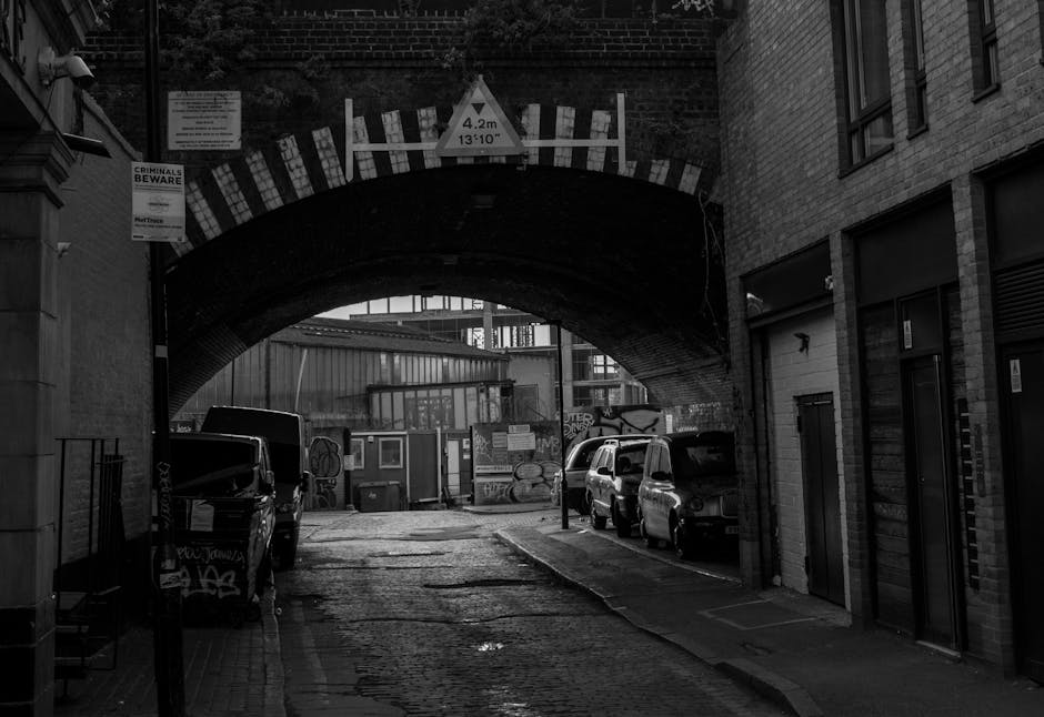 A narrow urban street in Stoke Newington E5, Hackney, shows a daytime scene with a wet asphalt road marked by a central white line, flanked on both sides by tall, mixed-use buildings with facades of brick, concrete, and metal cladding. On the left, there are modern black-framed glass windows and small shopfronts, while the right side features plain white and beige walls, some with windows and air conditioning units. Parked and moving vehicles, including a white van in the middle distance, occupy the street, which appears to have limited space for large rubbish removal vehicles to access directly. In the background, high-rise buildings and a cloudy sky are visible, contributing to the cityscape atmosphere. The setting suggests a typical city environment where alternative waste handling services might operate in tight or constrained access situations, as indicated by the closely spaced buildings and narrow street width; this aligns with the context of private rubbish collection and on-site clearance, as managed by Rubbish Clearance Hackney.