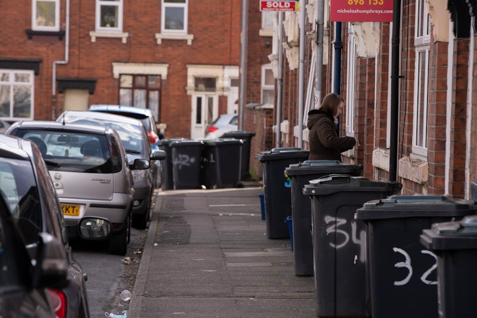 A residential street scene featuring a row of black wheelie bins with lid closures, lined up along the pavement beside brick terraced houses. The bins are positioned in close proximity, with some showing white painted numbers or markings, indicating designated rubbish collection points. A woman wearing a dark coat and hood stands next to one of the bins, seemingly placing or retrieving waste. The street is narrow, with several parked cars, including a silver hatchback in the foreground, along the side of the curb. The background shows more brick houses with white window frames, some with small front gardens, and a red sign hanging on one of the buildings. The environment appears to be an urban area prepared for or in the process of waste disposal, visually supporting private rubbish management services. The scene is captured in natural daylight with overcast sky, highlighting the textured brickwork and the orderly arrangement of refuse containers, consistent with independent rubbish clearance practices in residential neighbourhoods.