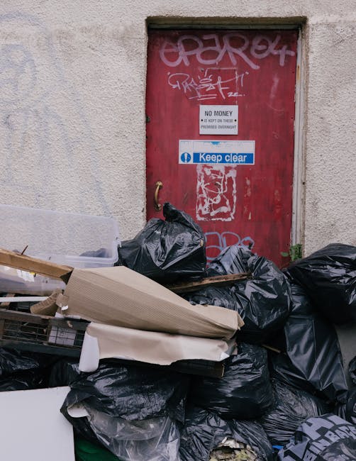 A close-up view of a utilitarian red metal door set into a beige, textured concrete wall, with graffiti tags in white and light pink spray paint decorating the upper portion of the door. Several black trash bags, some tightly tied and others loosely filled, are piled in front of the door, with a few leaning against the wall and obscuring the lower part of the door frame. Among the rubbish are loose cardboard pieces, some flattened and others rolled, as well as a cluttered collection of miscellaneous waste, suggesting an accumulation of discarded household or commercial waste. A small sign on the door displays a white background with black text stating 'NO MONEY' and additional information, accompanied by a blue and white sticker indicating 'Keep clear.' The overall scene appears to be in an urban alleyway or rear access area, with natural lighting and shadows emphasizing the textures of the trash and wall surface, illustrating a scene aligned with private waste collection or rubbish removal services, as managed by Rubbish Clearance Hackney.